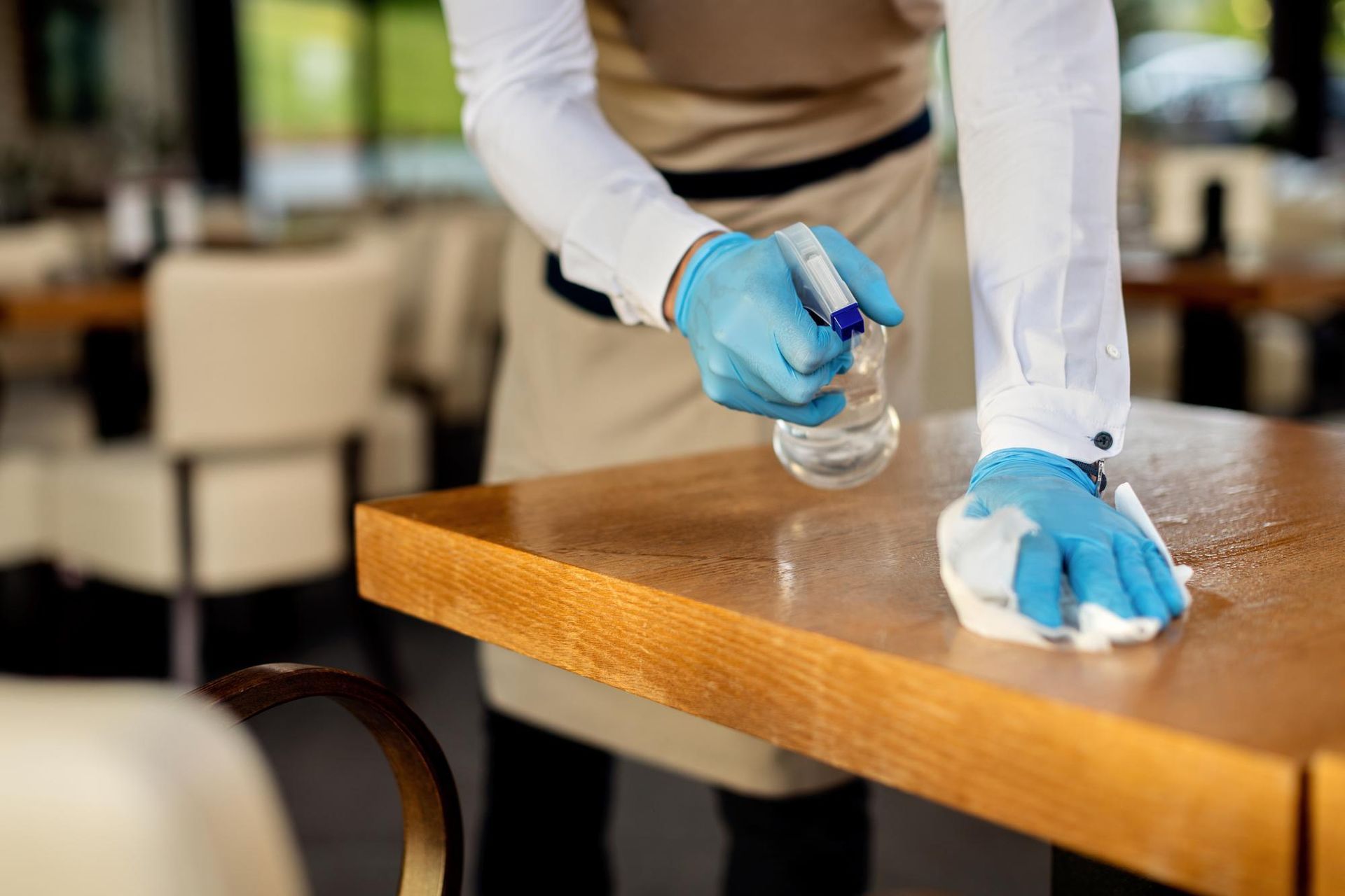 A man wearing blue gloves is cleaning a wooden table in a restaurant.